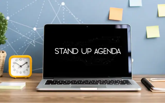 A laptop on a desk showing the words Stand Up Agenda, next to a yellow alarm clock and sticky notes on the wall
